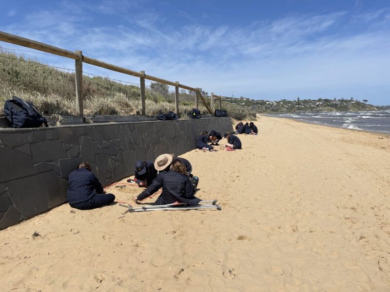 People sit along a seawall on a sandy beach, leaning over and engaging in an activity. Backpacks are placed nearby, and the sea is visible to the right.
