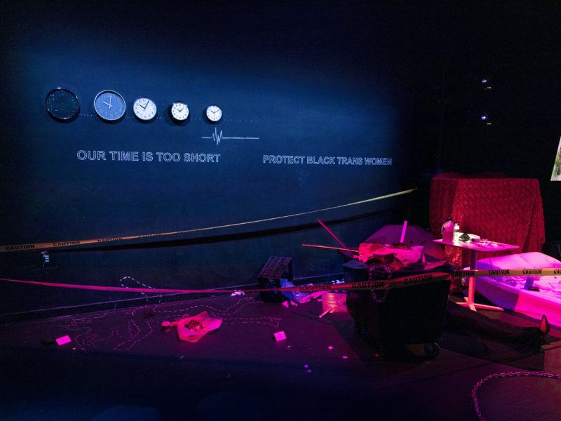 Multiple installations by Lexii Fox at Performance Space, white text on the wall reads "Our time is too short, protect Black Trans Women." The text is beneath five clocks mounted on the wall. On the floor is a chalk body outline, surrounded by police caution tape and assorted items.