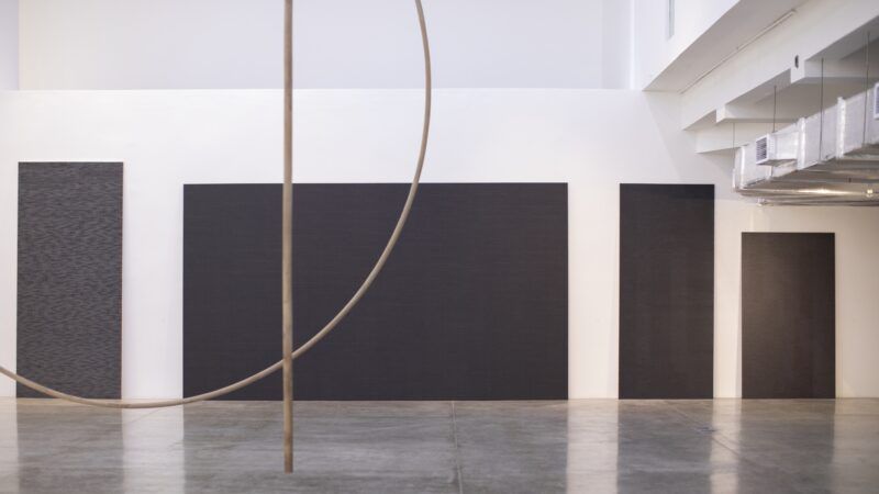 Four large, rectangular black panels are mounted on a white wall in a gallery space with polished concrete floors and a curved sculpture in the foreground.