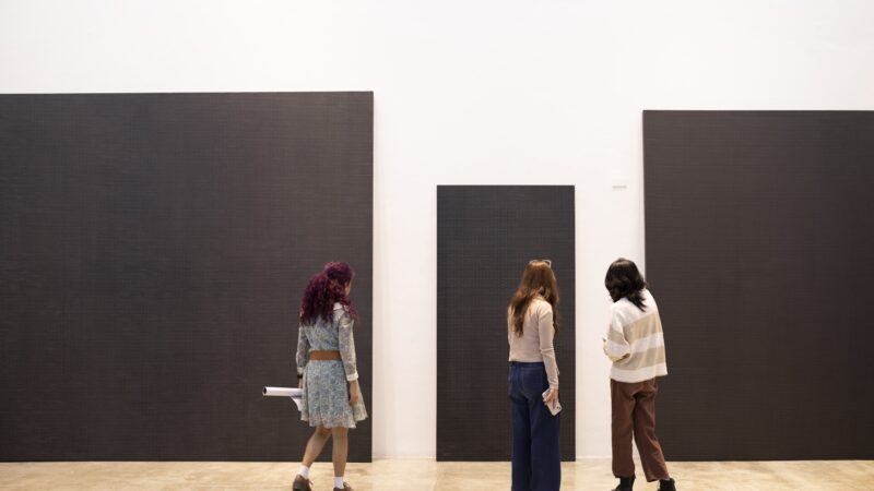 Three people stand in front of large, minimalist black rectangular artworks displayed on a white gallery wall.