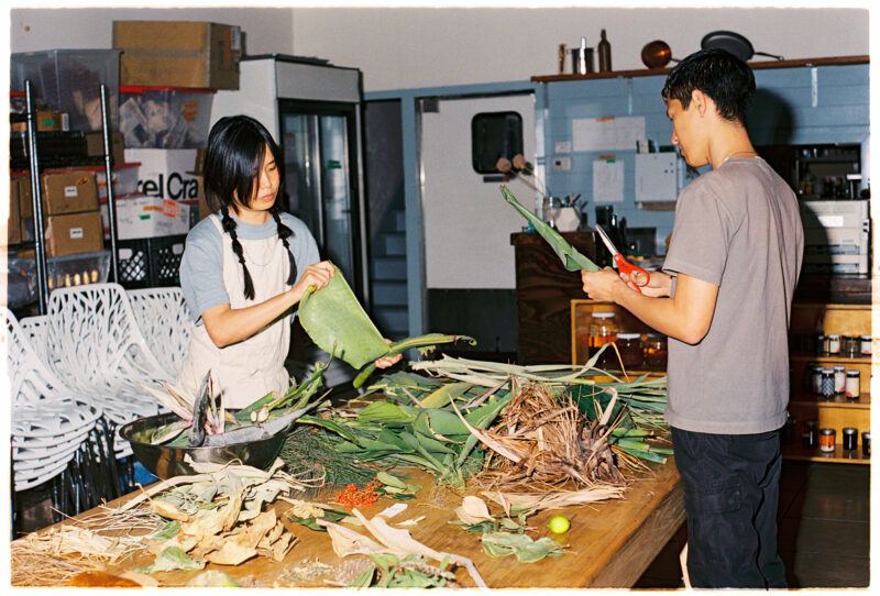 Two people work at a wooden table filled with various types of leaves.