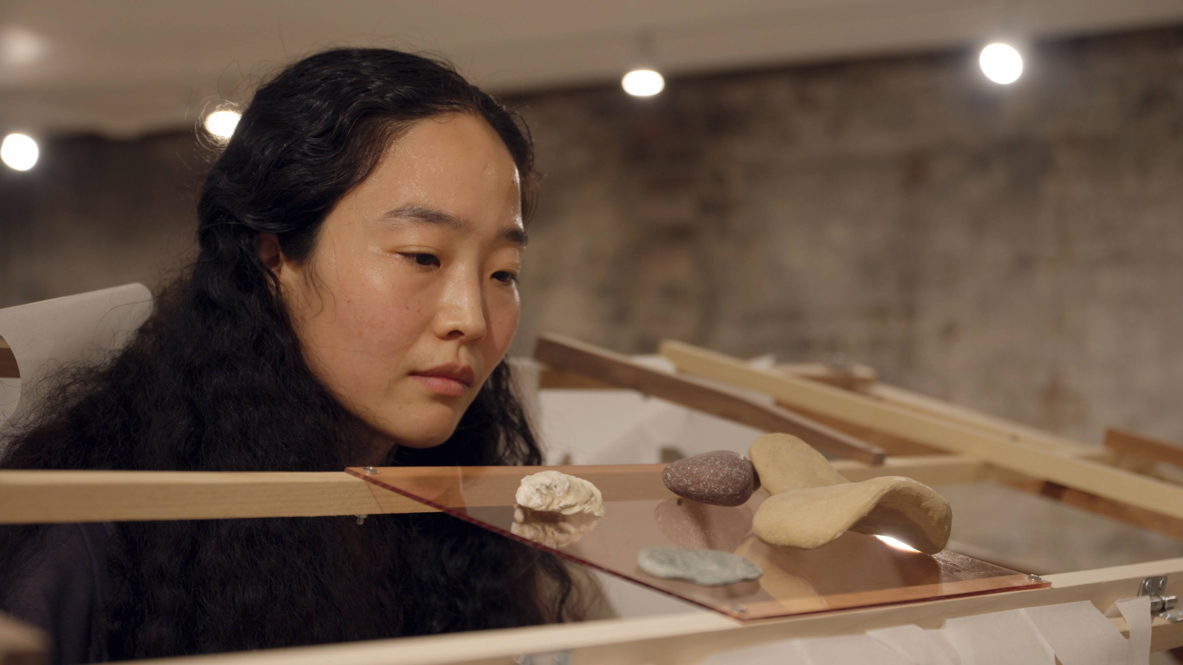 Hyeree Ro with long dark hair closely observes small sculpted objects displayed on a transparent surface supported by a wooden frame.