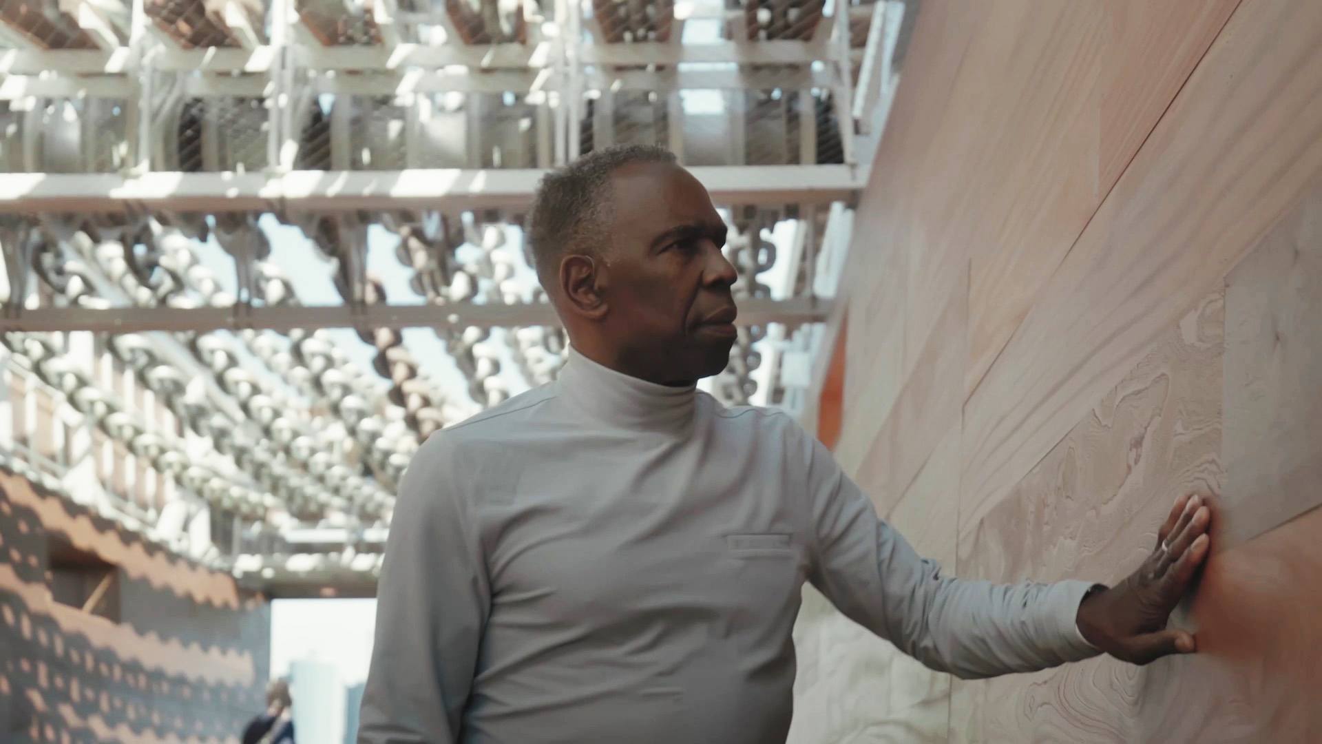 Charles Gaines standing inside his sculpture "Moving Chains." He places his hand on the wall next to him, and his looking up at the oversized chains above.