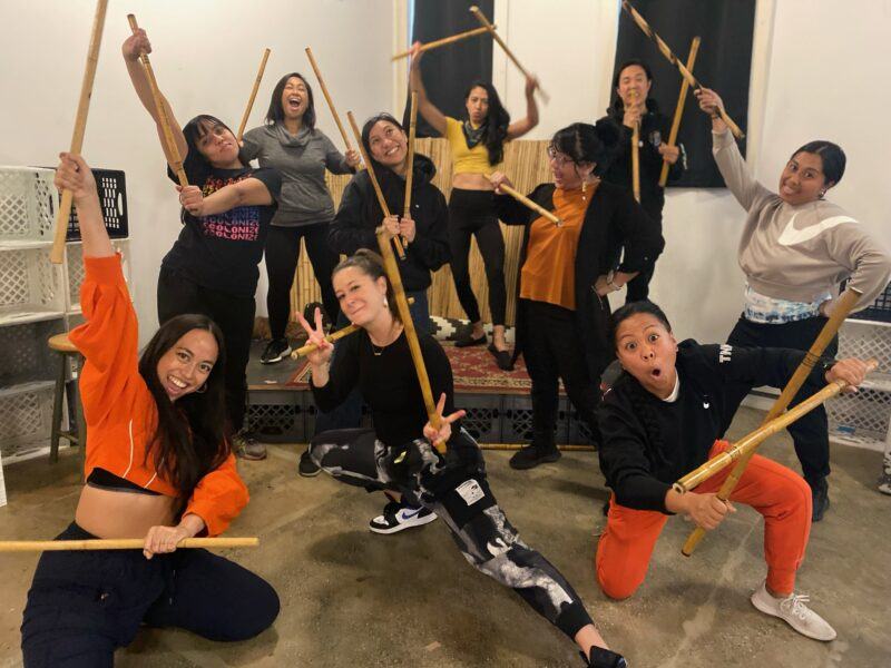 A group of people pose enthusiastically indoors holding wooden sticks, appearing to practice or perform a martial arts or stick-fighting routine.
