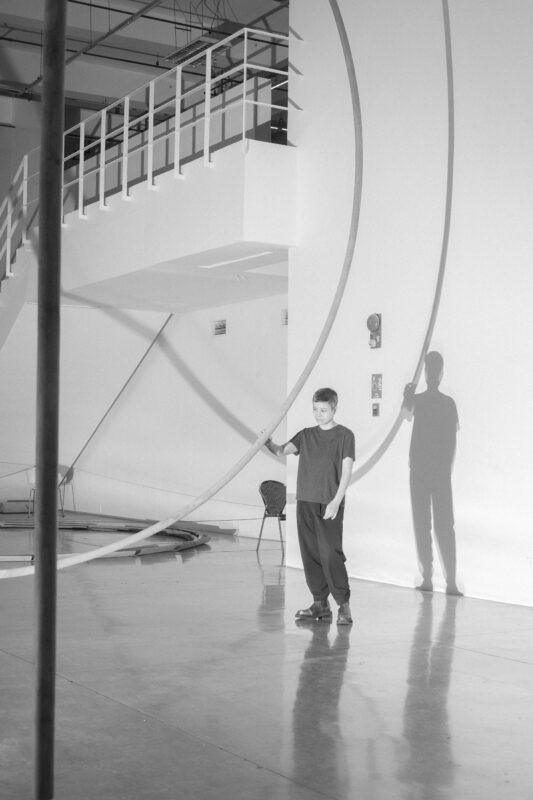 Black and white image of Maria Taniguchi standing indoors holding onto a long curved pole, casting a shadow on a white wall; industrial stairs are visible in the background.