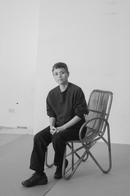 Black and white image of Maria Taniguchi sits on a wooden chair in a minimalist room, wearing a dark sweater and pants.