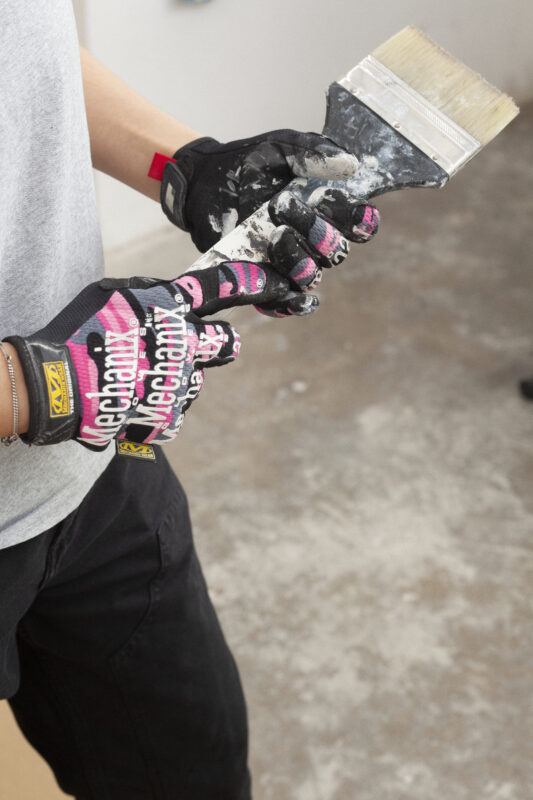 Hands wearing gloves with "Mechanix" on them, holding a paintbrush with white paint on it, standing on a concrete floor.