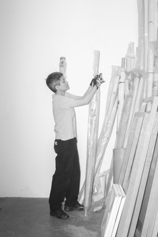 Black and white image of Maria Taniguchi wearing gloves stands in a room, handling a large rolled object among several wrapped objects and frames leaning against a white wall.