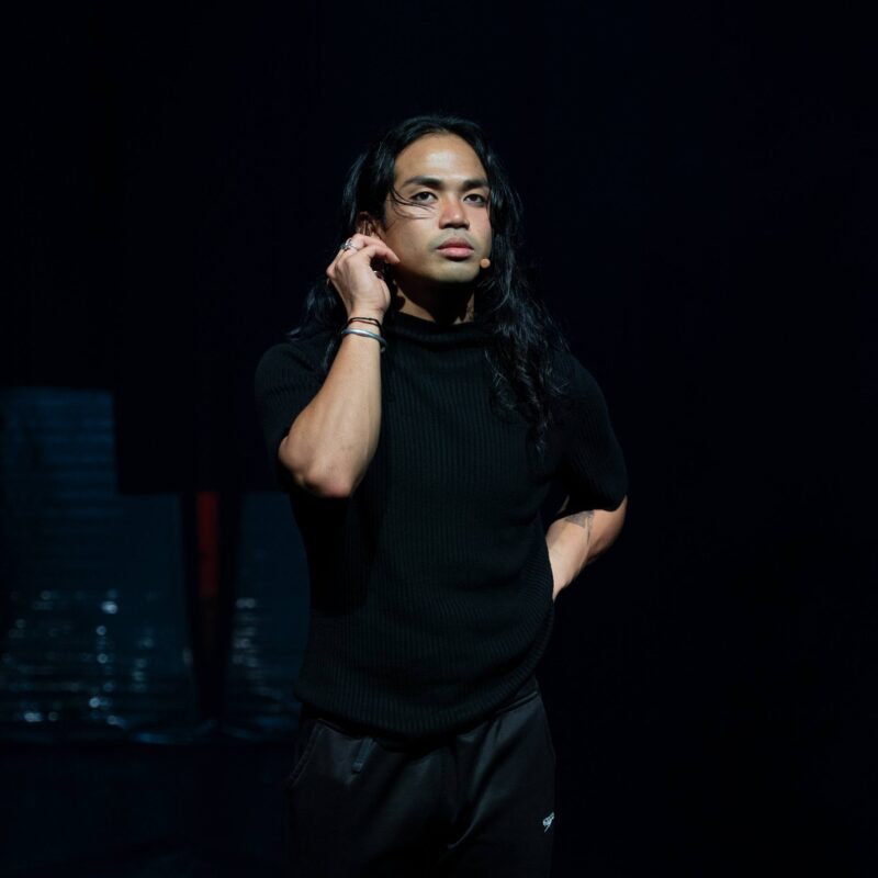 Joshua Serafin with long hair and face paint stands on a dimly lit stage, wearing a black shirt and pants, with one hand touching their ear and the other behind their back.