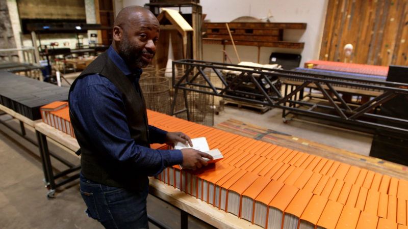 Theaster Gates standing in his studio assembling orange bound books on a table, spines up.