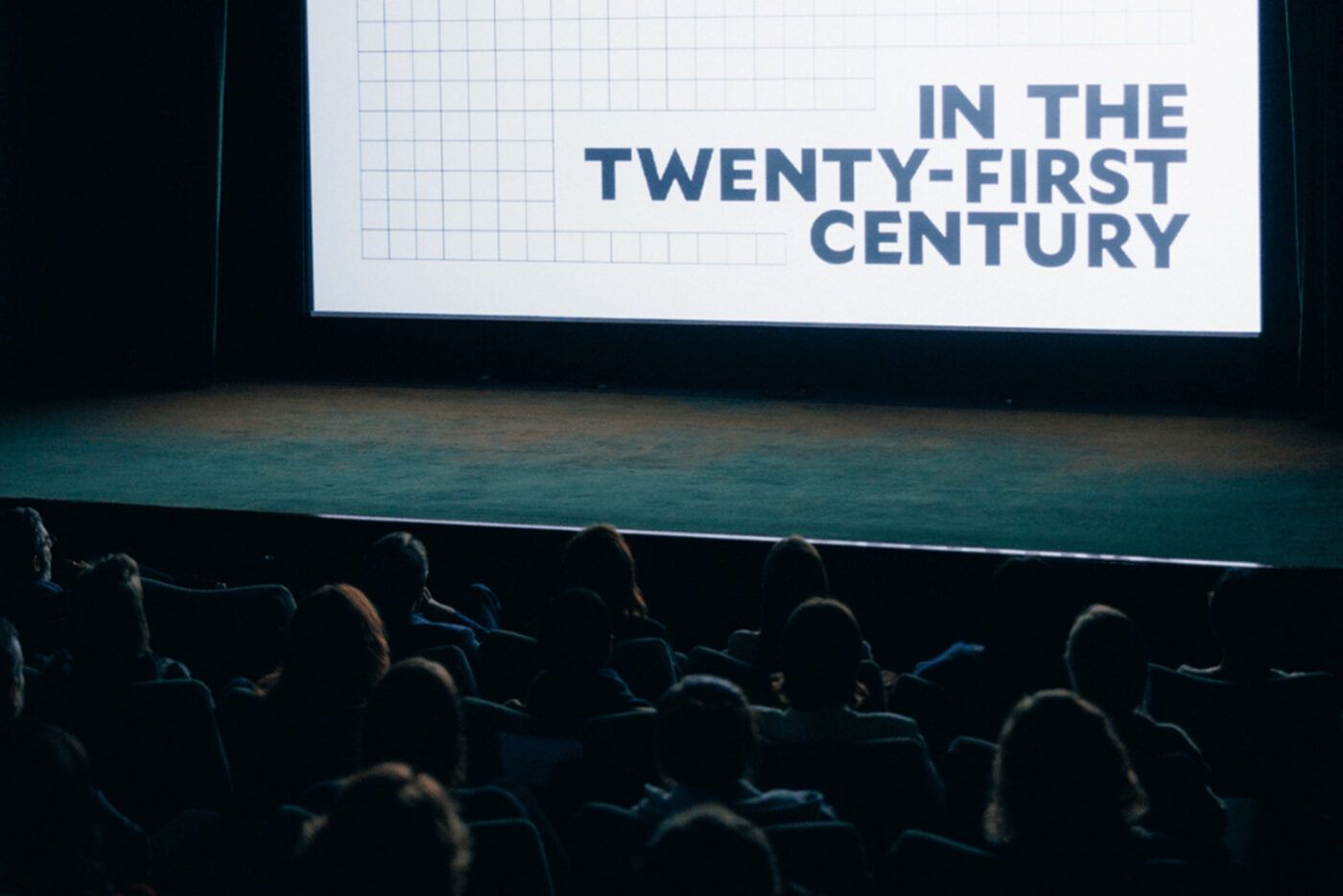 Audience seated in a dark theater, watching a large screen displaying the words "IN THE TWENTY-FIRST CENTURY" in bold text.