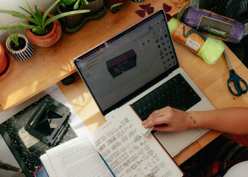 A person uses a laptop with a 3D modeling program open, referencing a book with patterns. Yarn, scissors, and potted plants are on the wooden desk.