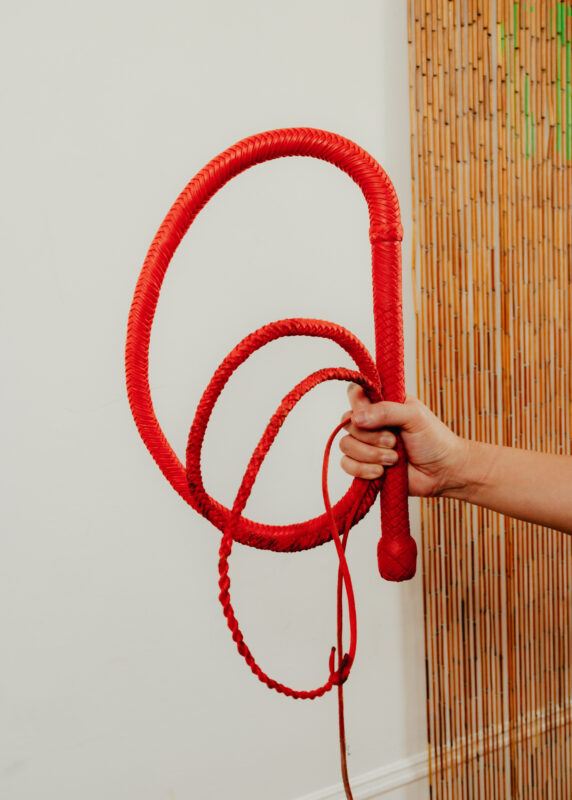 A hand holding a coiled red whip in front of a white wall and bamboo curtain.
