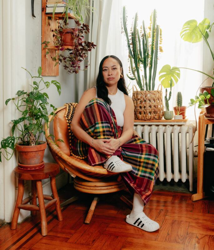 Caroline Garcia in a white sleeveless top and plaid skirt sits on a chair surrounded by various potted plants in a sunlit room.