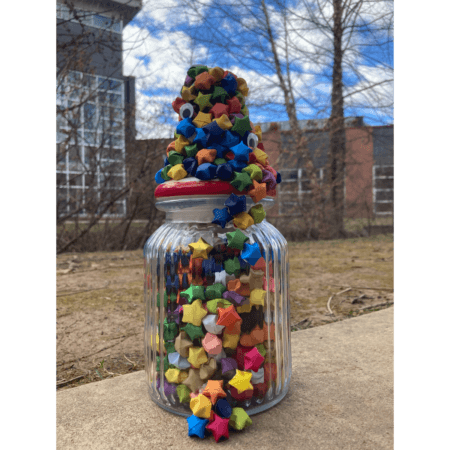 A glass jar filled with colorful paper stars sits outside on a concrete surface, stars with googly eyes are stacked and spilling out of the top.